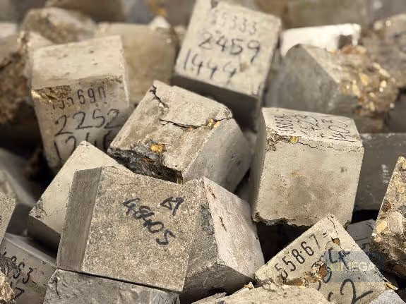 Broken grey concrete cubes and blocks thrown at many angles into a metal waste skip—handwritten numbers on each surface. The cubes are cracked and chipped, conveying a sense of decay and age.