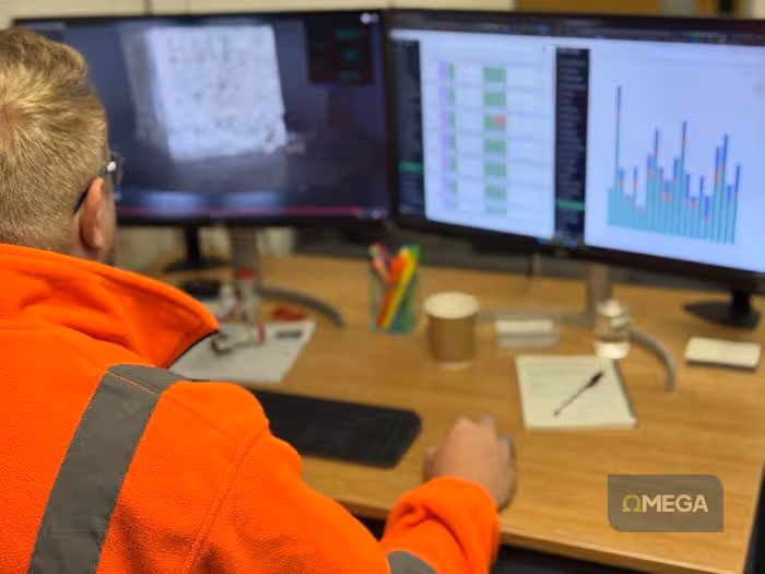 A person in a bright orange jacket analyses a test on dual monitors. The left screen shows a video preview, the right features a bar chart and a table. Desk items include pens and a coffee cup. The setting is an organised, focused workspace.