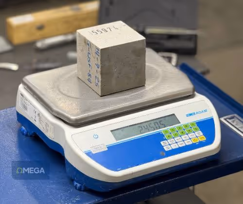 A concrete block with numbered markings is placed on a calibrated digital scale, which displays the air weight. The scale is on a blue platform in an industrial setting.