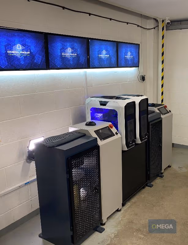 A room with three high-tech machines, blue flatscreens above on the wall displaying "The Omega Group" branding, and illuminated with a bright, modern industrial vibe.