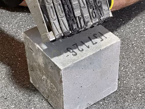 A close-up of a metal stamp pressing the unique sample ID number onto the surface of a grey concrete block.
