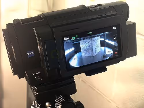 A high-definition video camera on a tripod films a cube test under bright lights. Its screen shows the recording in progress.