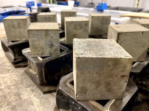 Nine grey concrete cubes resting on top of steel cube moulds in rows on a work surface in a lab. One cube is in clear focus. 
