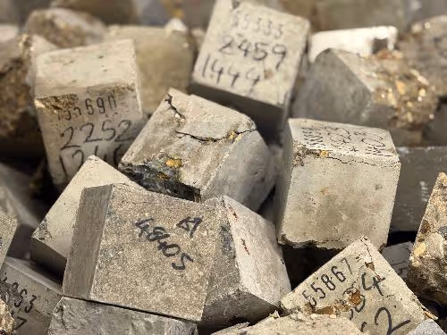 A pile of broken and cracked grey concrete cubes in a waste skip. Each block is labelled with a sample ID and density data.