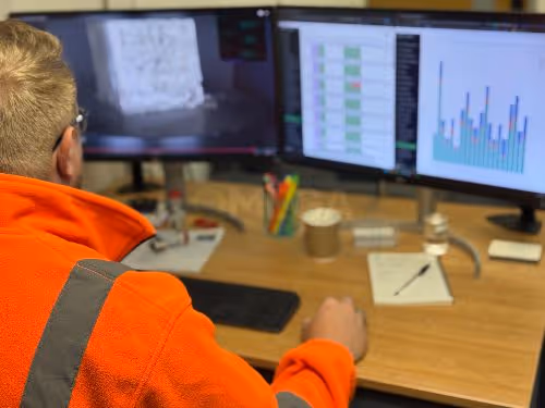 A person wearing an orange high-vis jacket sits at a desk. They review a bar chart, a video, and test results on two monitors.