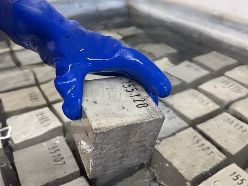 A gloved hand holds a numbered concrete cube over a water curing tank, surrounded by other similar cubes.