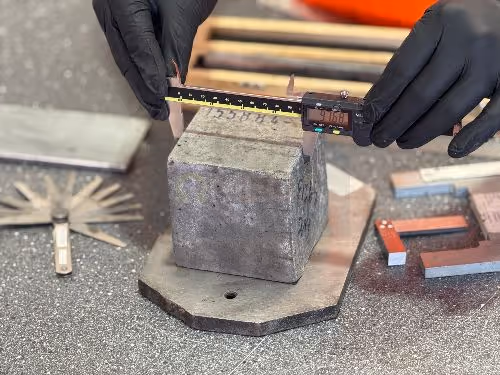 A person wearing black gloves uses a digital calliper to measure the size of a concrete cube on a workbench.