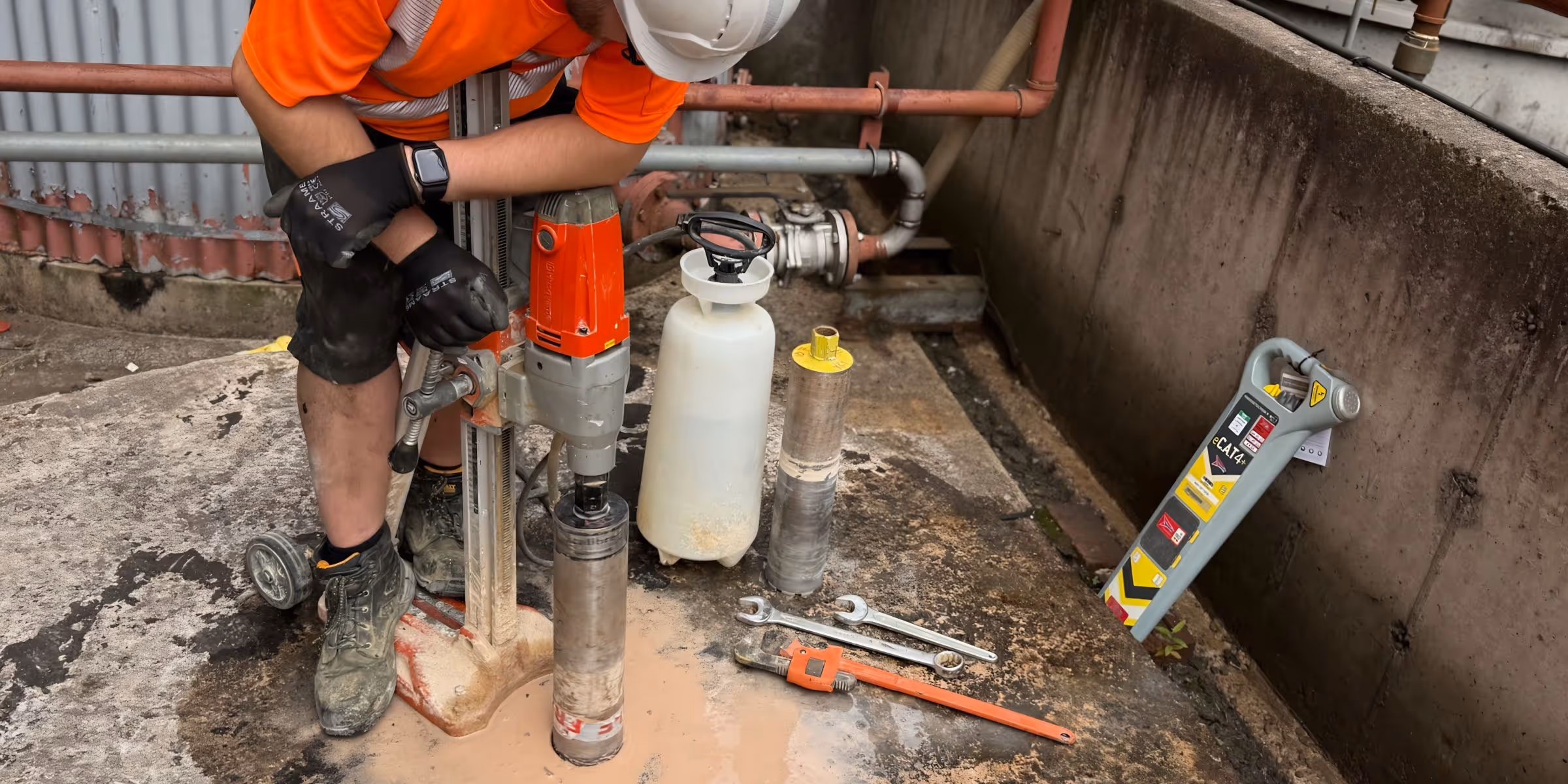 A technician is working with a core drilling machine on a concrete surface. This demonstrates the process of diamond drilling and concrete coring. Nearby, you can see tools, a water container, and a CAT scanner.