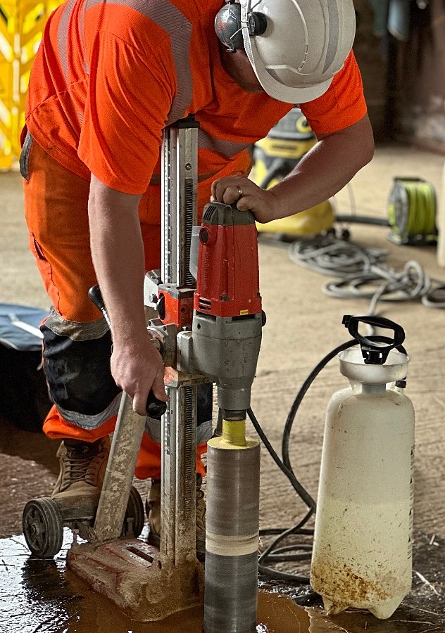 A construction worker in a white helmet and orange safety gear operates a concrete coring drill. Nearby, a spray bottle and tangled cables lie on the floor.