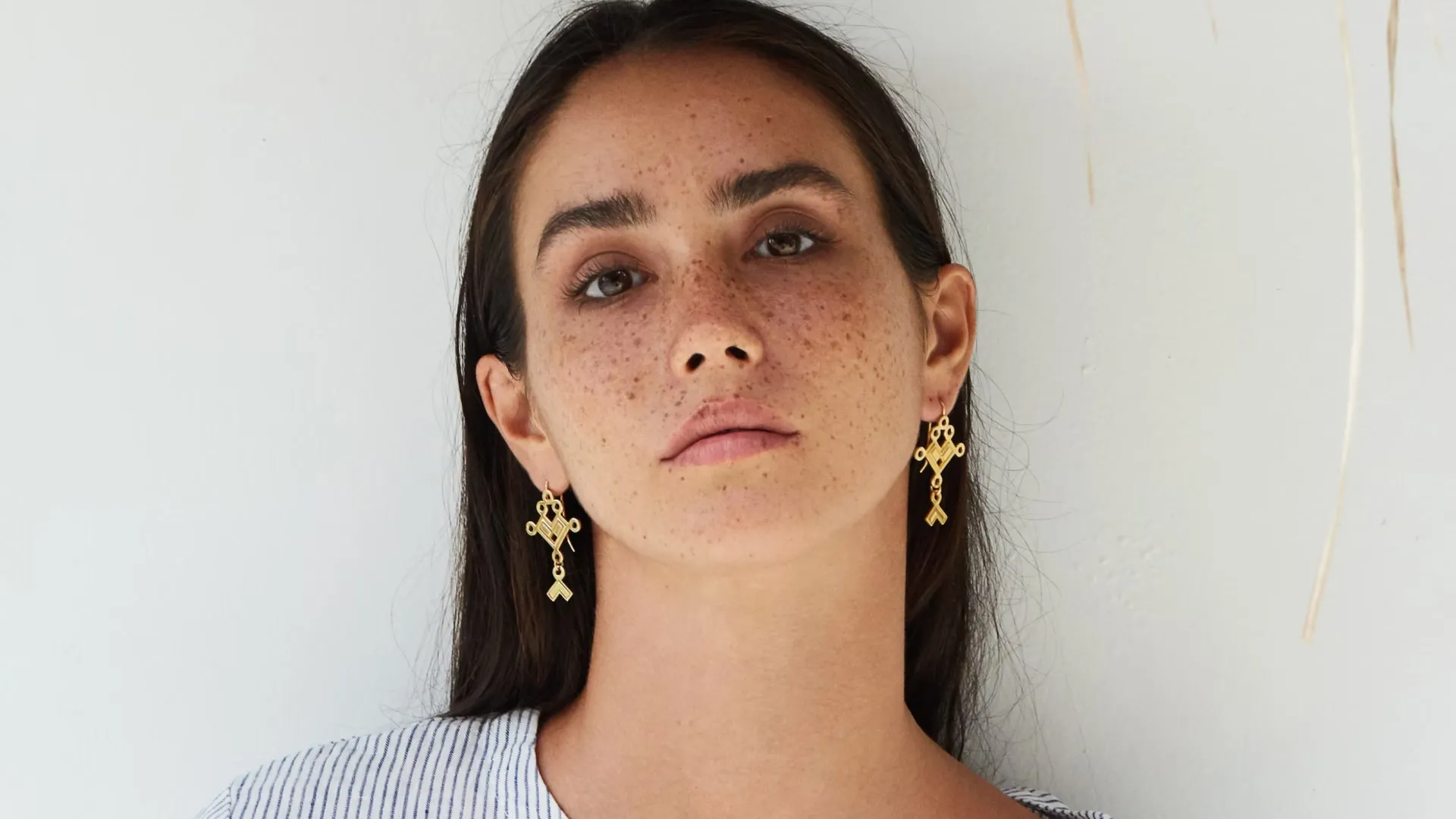 Close-up of a woman with freckles wearing gold decorative earrings and a striped top against a white background.