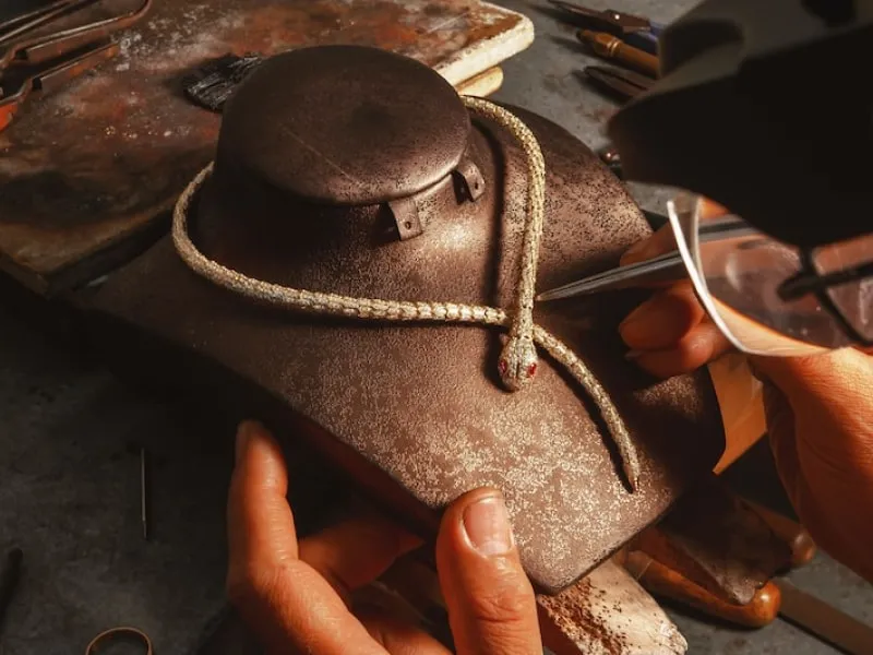 Hands crafting a braided rope snake with red eyes on a brown textured display stand.