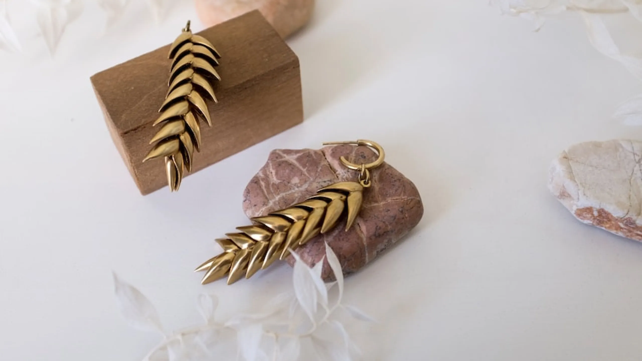 Pair of gold leaf-shaped earrings displayed on brown wooden block and pink stone.
