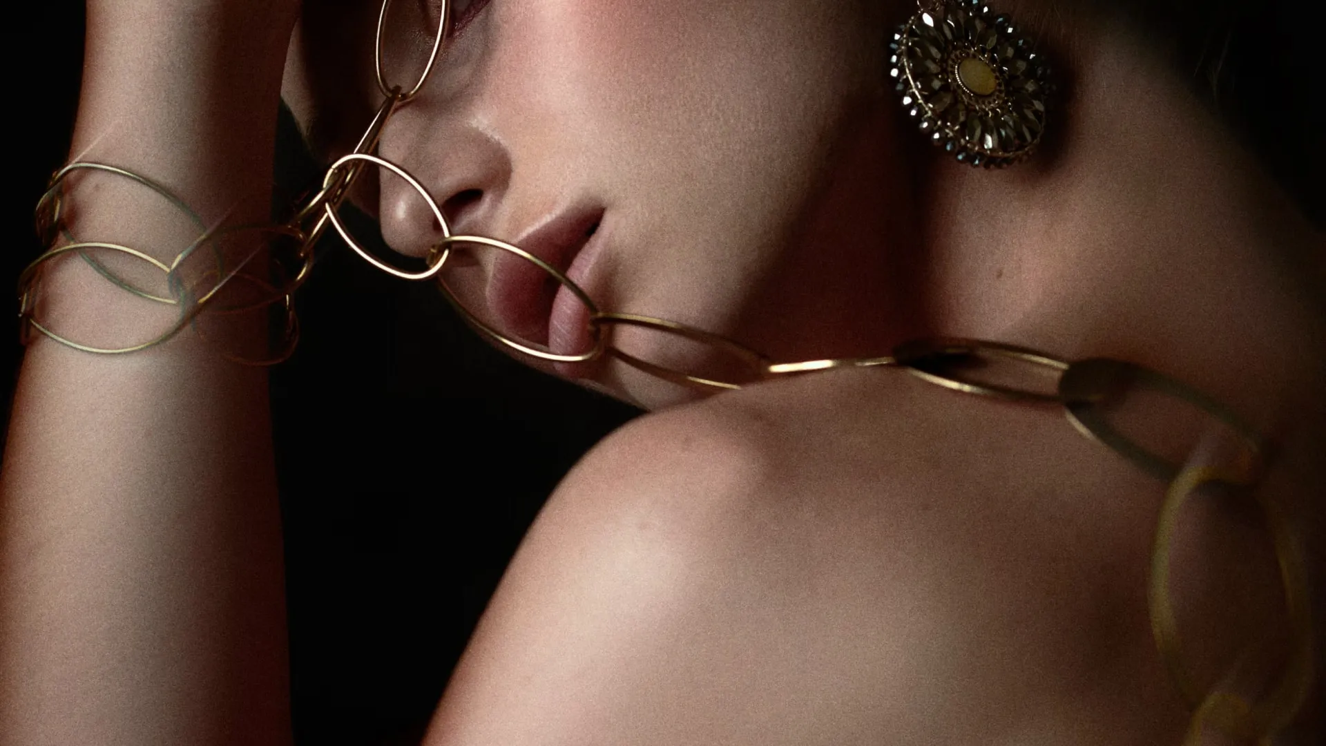 Close-up of a woman with gold chain jewelry on her arm and soft light on her face and shoulder.