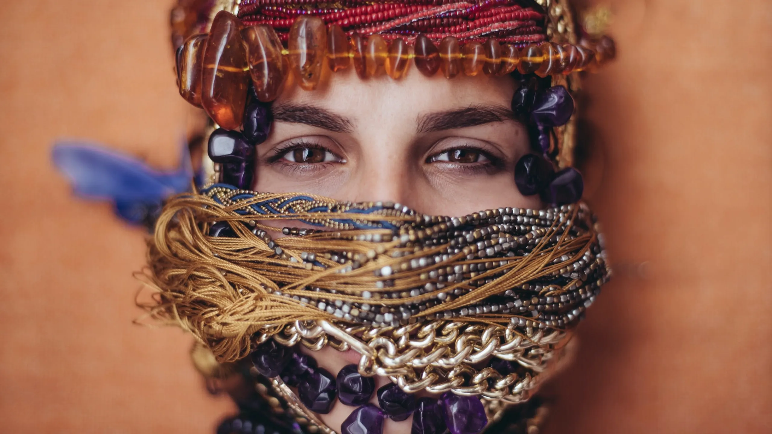 Person's face mostly covered with gold chains, beads, and fabric, revealing only brown eyes.