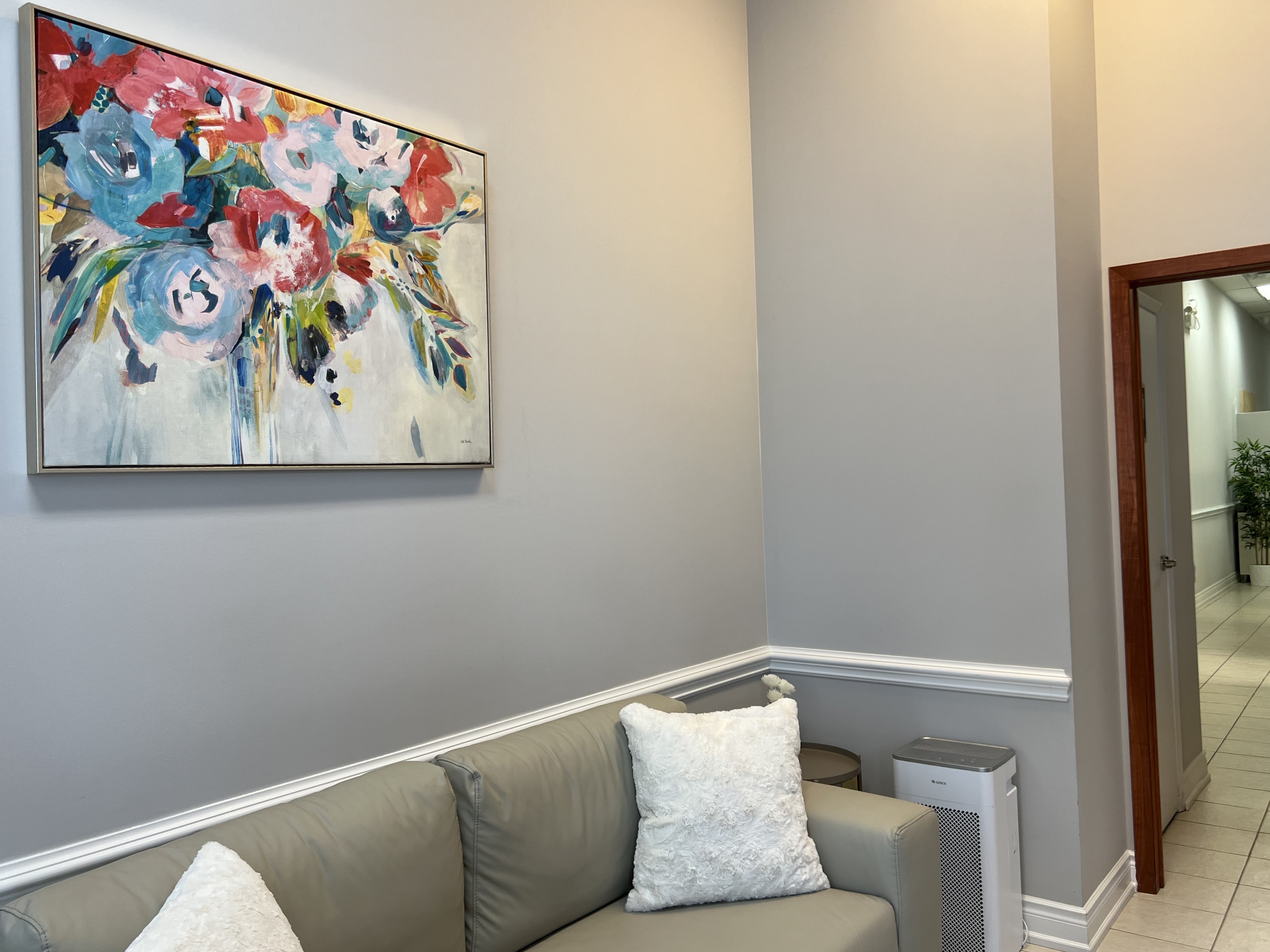Modern waiting area with gray couch, white pillows, a floral painting, and air purifier near hallway.