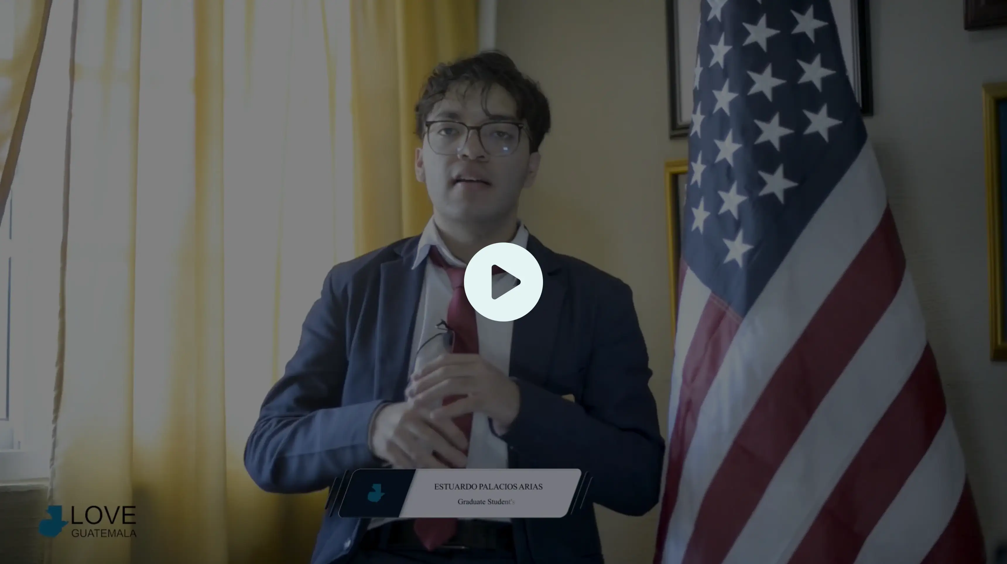 Young man in glasses wearing a suit and red tie sitting beside an American flag with yellow curtains in the background.