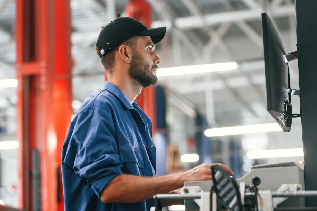Worker in blue uniform and cap operating a machine and looking at a monitor in an industrial setting.