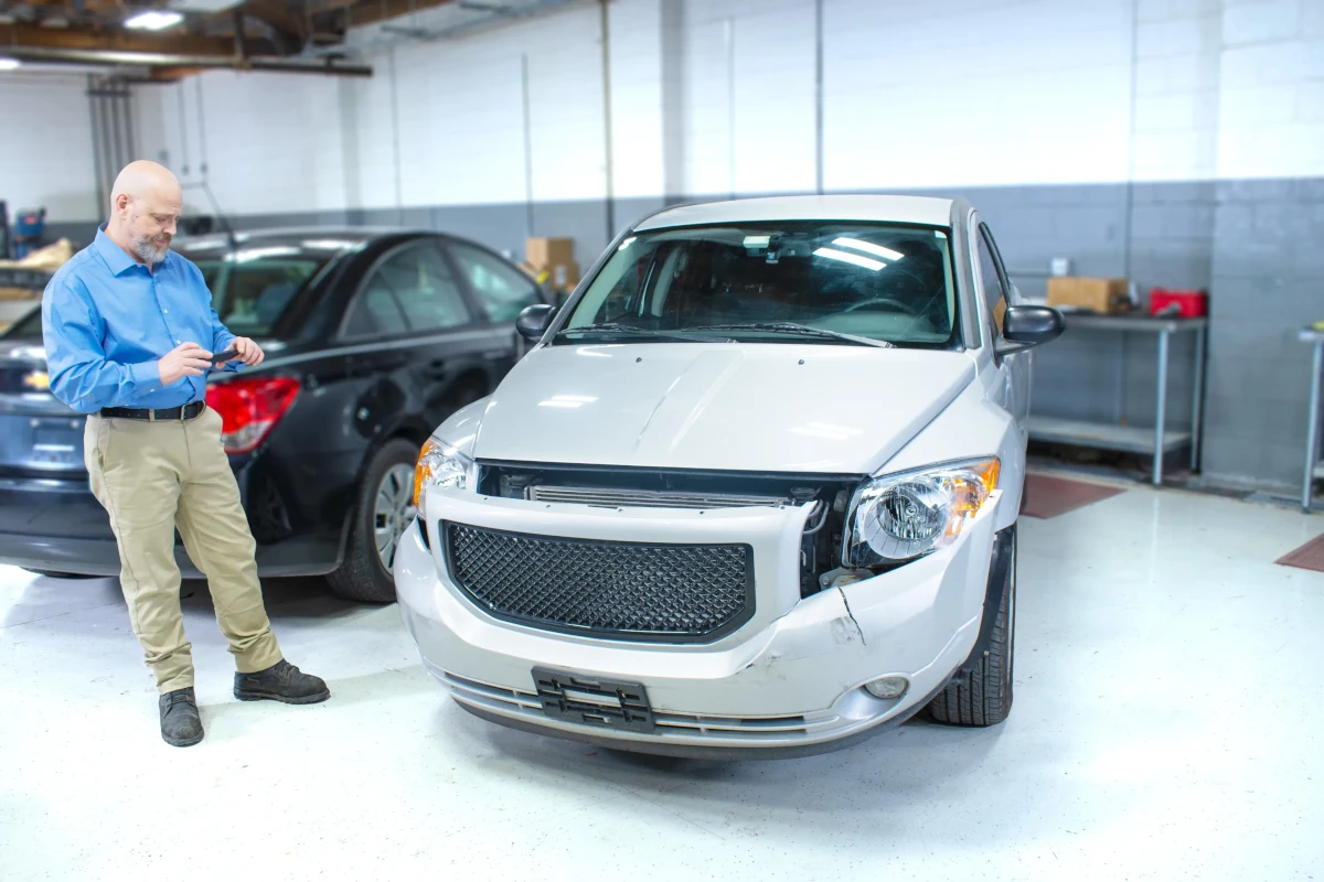 Man inspecting damage on front bumper and headlight of silver car in garage.
