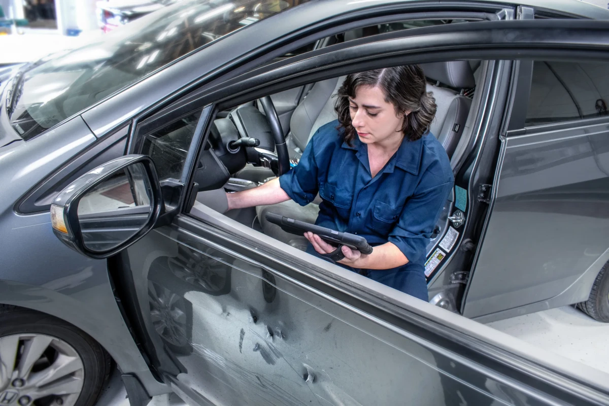 Woman in blue uniform inspecting damage on car door using a diagnostic tablet inside the vehicle.