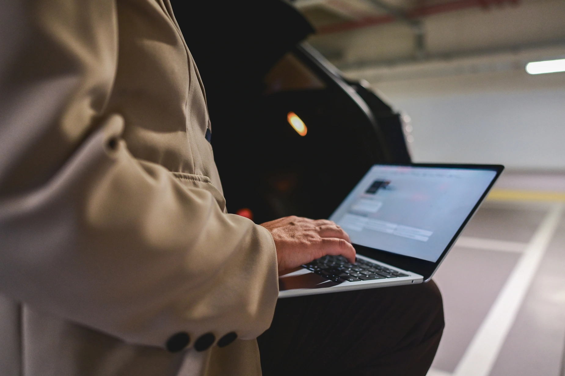 Person wearing a beige coat typing on a laptop in a dimly lit parking garage with a black car in the background.