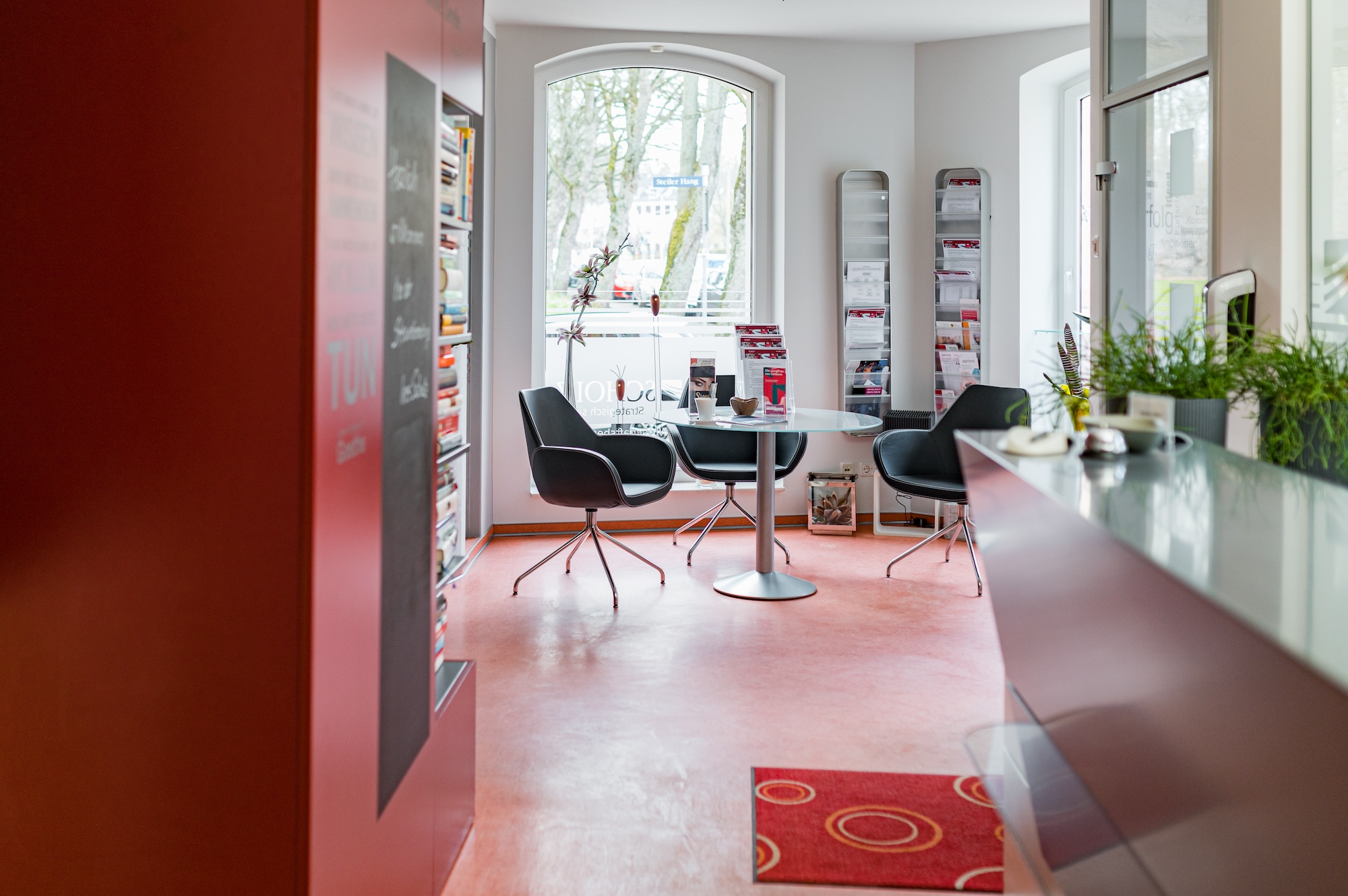 Heller Wartebereich mit drei schwarzen Drehstühlen um einen runden Glastisch, großem Fenster im Hintergrund und Literaturständern auf beiden Seiten.
