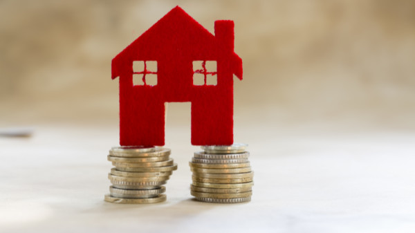 Rote Filzhaus-Figur steht auf zwei Münzstapeln auf einer beigen Oberfläche.