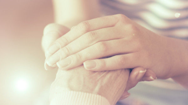 Junge Person hält sanft die Hand einer älteren Person mit hellem Sonnenlicht im Hintergrund.