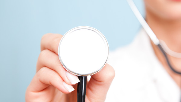 Nahaufnahme einer Hand, welche ein Stethoskop in Richtung der Kamera hält, vor einem hellblauen Hintergund.