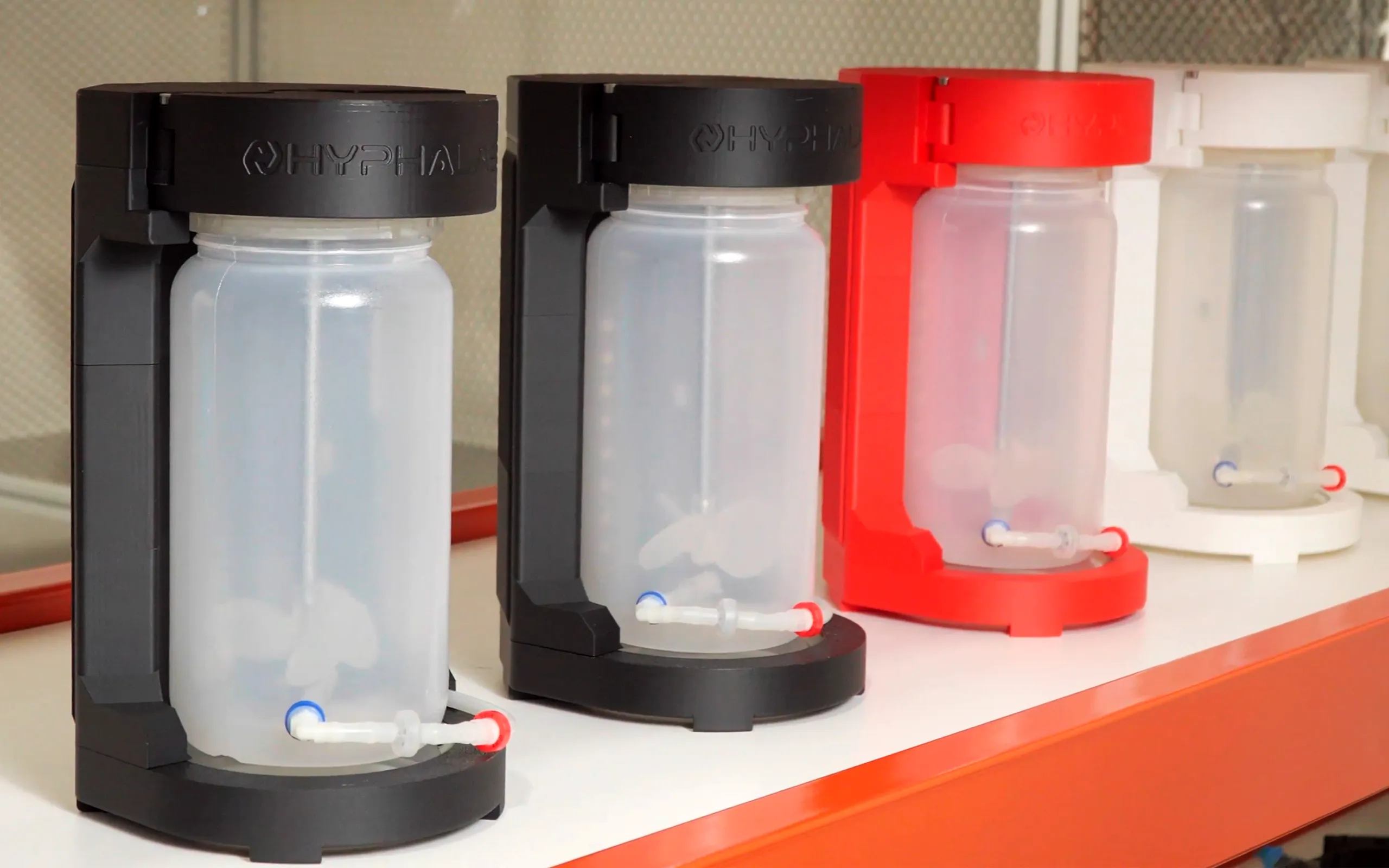 Line of Hypha Labs units—two black, one red, and a white model in the back—each holding an empty translucent jar with tubing at the base, arranged on a workbench.