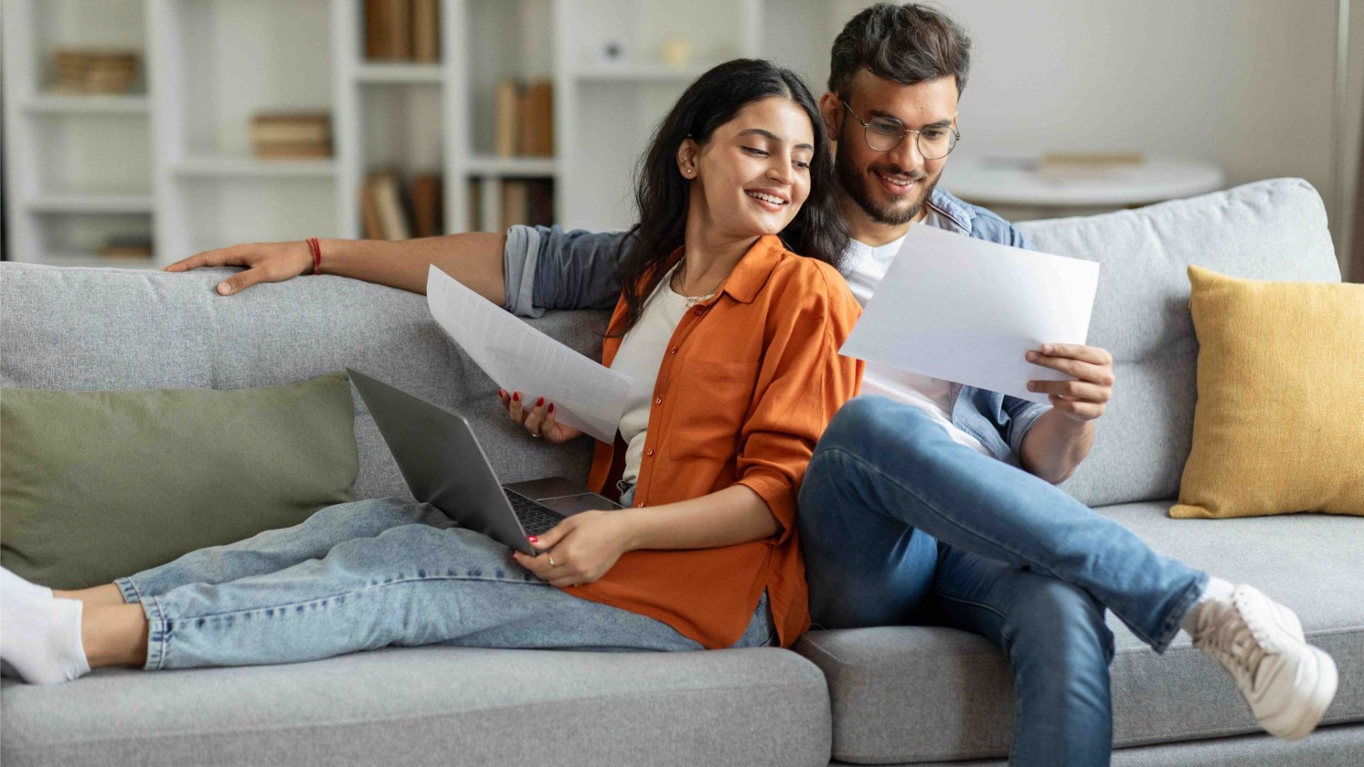 Junges Paar sitzt lächelnd auf dem Sofa, prüft gemeinsam Finanzierungsunterlagen