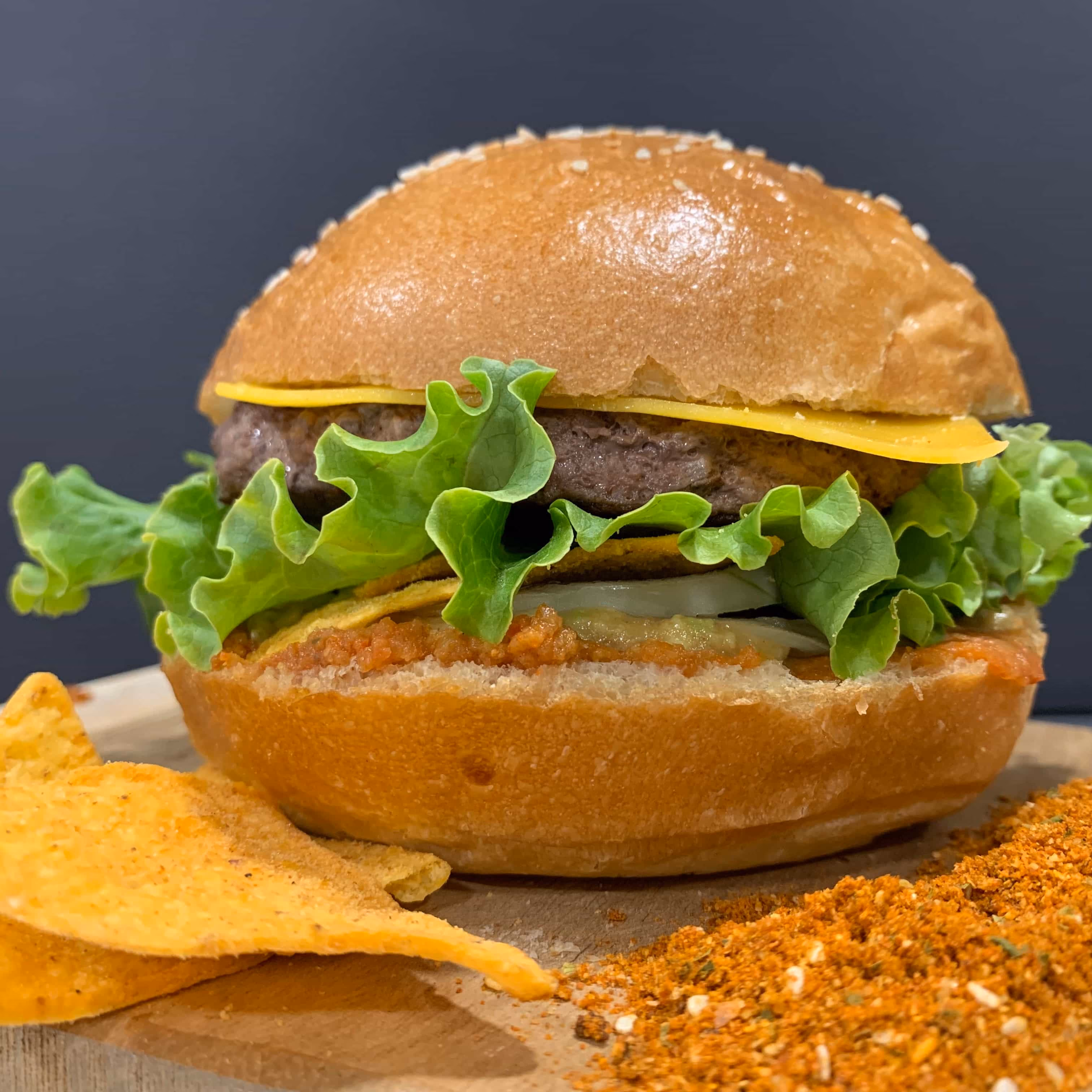 Un hamburger avec une boule de pain brun à graines de sésame, laitue verte, tranche de fromage, steak haché, et sauce rouge, accompagné de chips triangulaires et d'épices moulues à côté, sur une planche en bois.