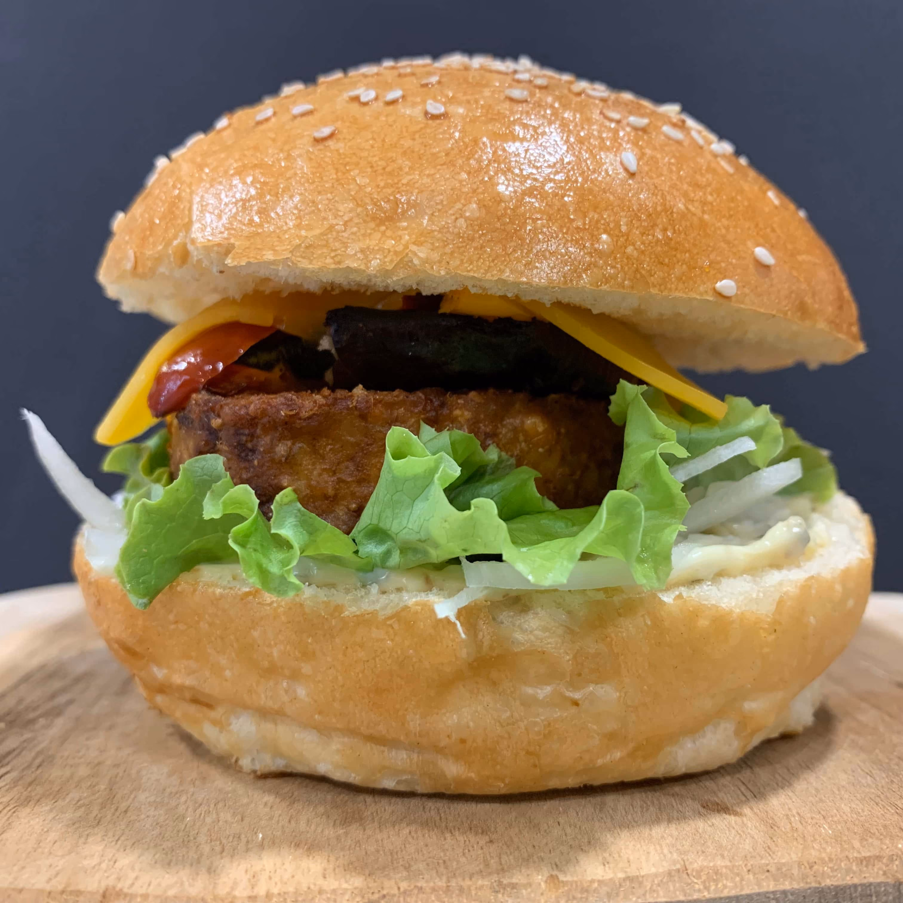 Burger végétarien avec une tranche de cheddar, une galette croustillante, de la laitue, des oignons émincés et une sauce blanche dans un petit pain à graines de sésame, posé sur une planche en bois avec un fond sombre.