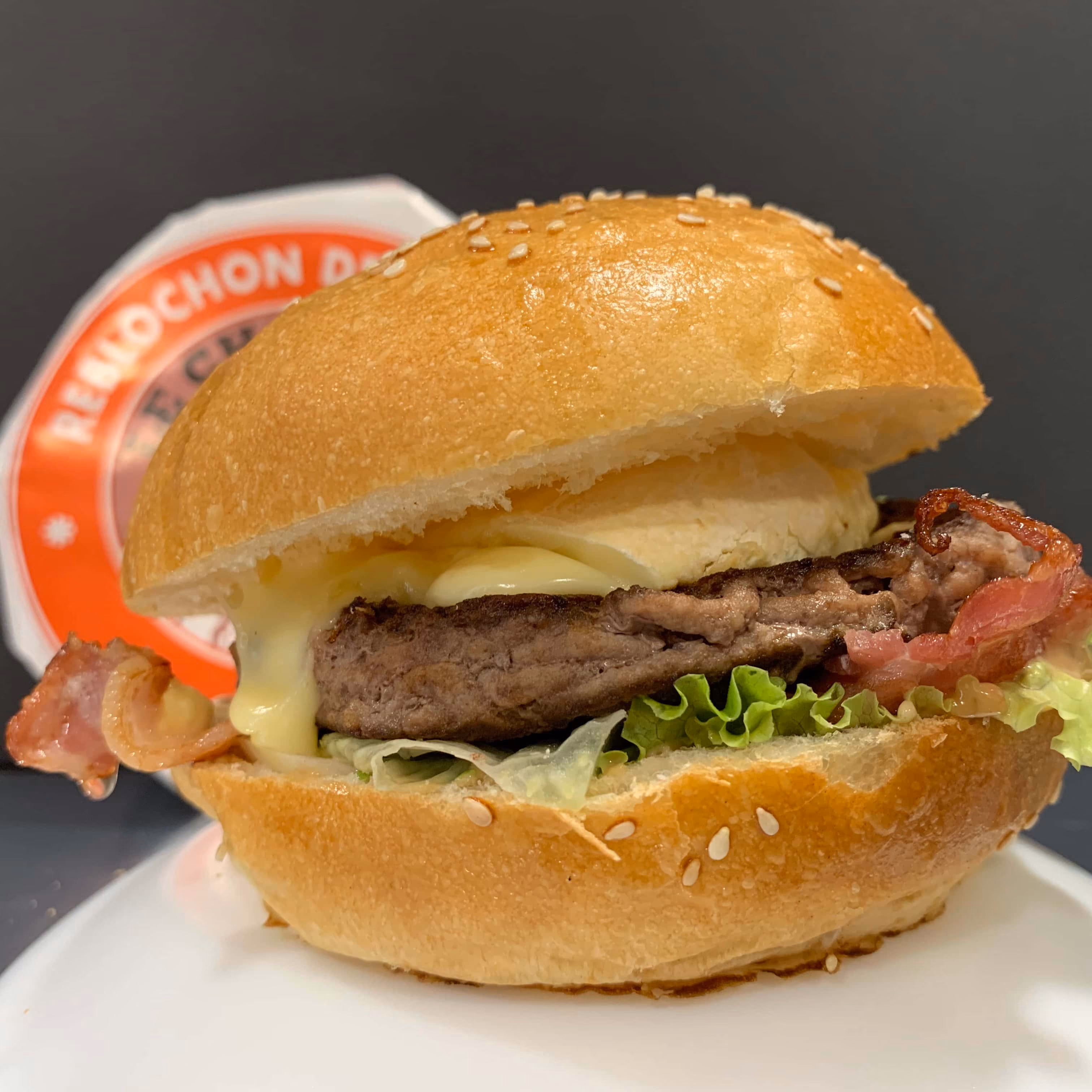 Burger avec un steak grillé, bacon, laitue, et fromage fondu dans un petit pain au sésame, posé sur une assiette blanche avec un logo rouge en arrière-plan.