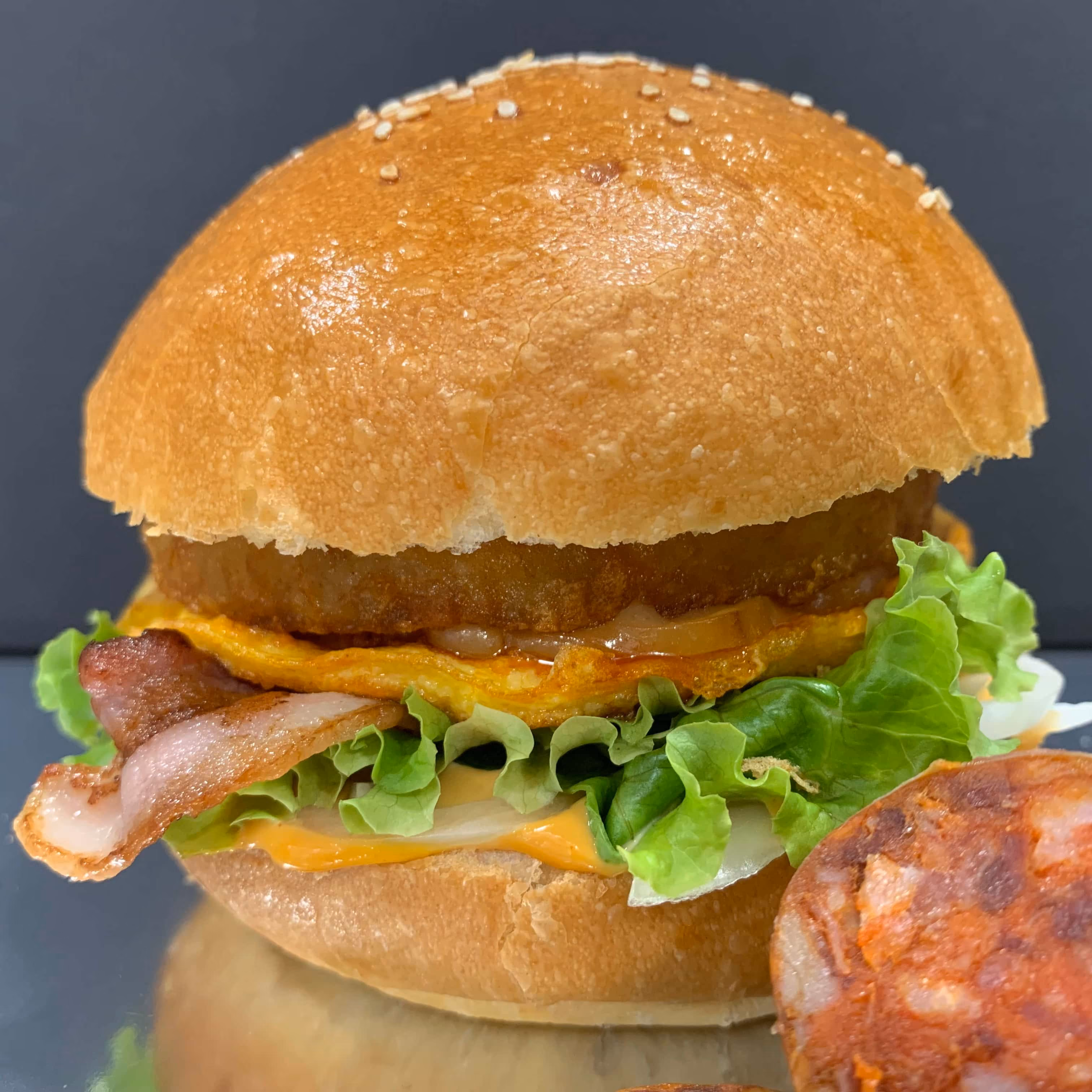 Un hamburger avec un pain à graines de sésame, de la laitue verte, une tranche de bacon croustillant, un œuf frit, une tranche de fromage fondu, et un morceau de viande ronde, sur un fond sombre.