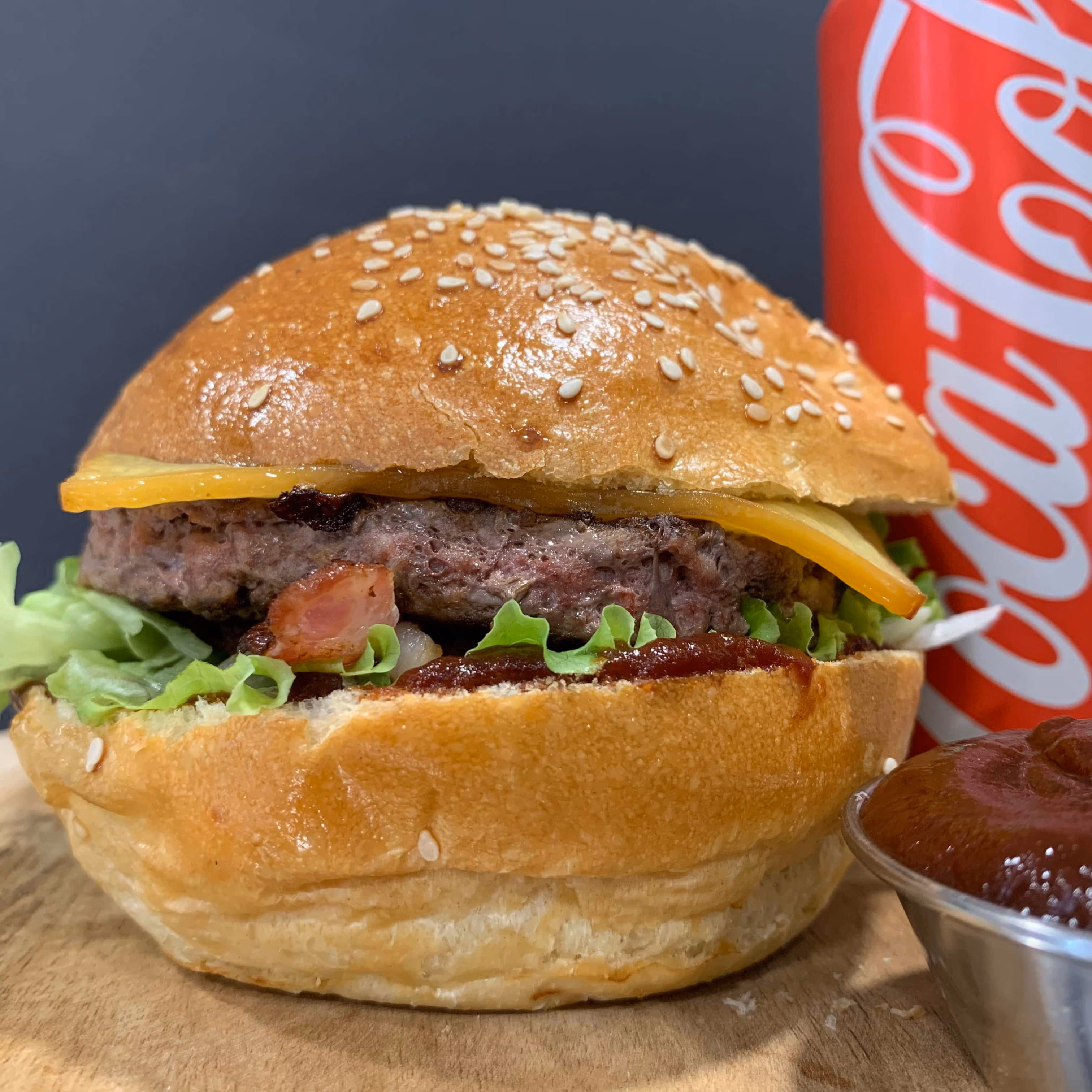 Un hamburger avec un pain aux graines de sésame, une tranche de fromage cheddar, de la laitue, du bacon et une sauce barbecue, accompagné d'une canette de Coca-Cola rouge et d'une petite coupelle de sauce barbecue sur une surface en bois.