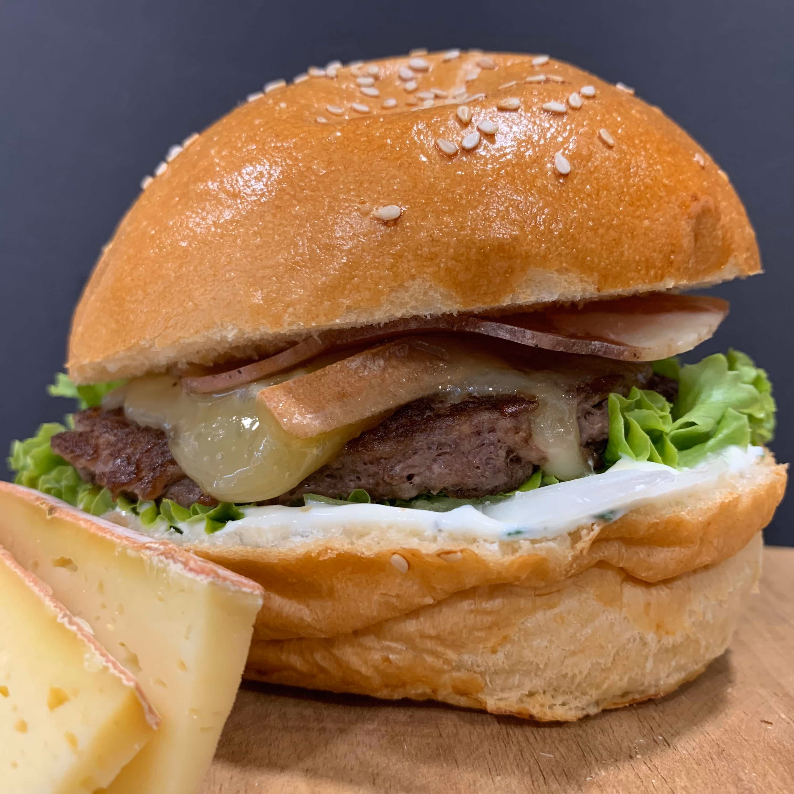 Un hamburger avec un pain brioché parsemé de graines de sésame, une tranche de fromage fondu, de la viande hachée, de la laitue verte, une sauce blanche, et des tranches fines de charcuterie, posé sur une planche en bois avec deux morceaux de fromage à côté.