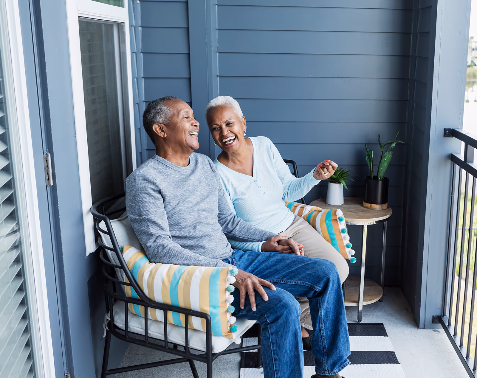 Older couple smiling and sitting together on a balcony, holding hands and looking at each other.
