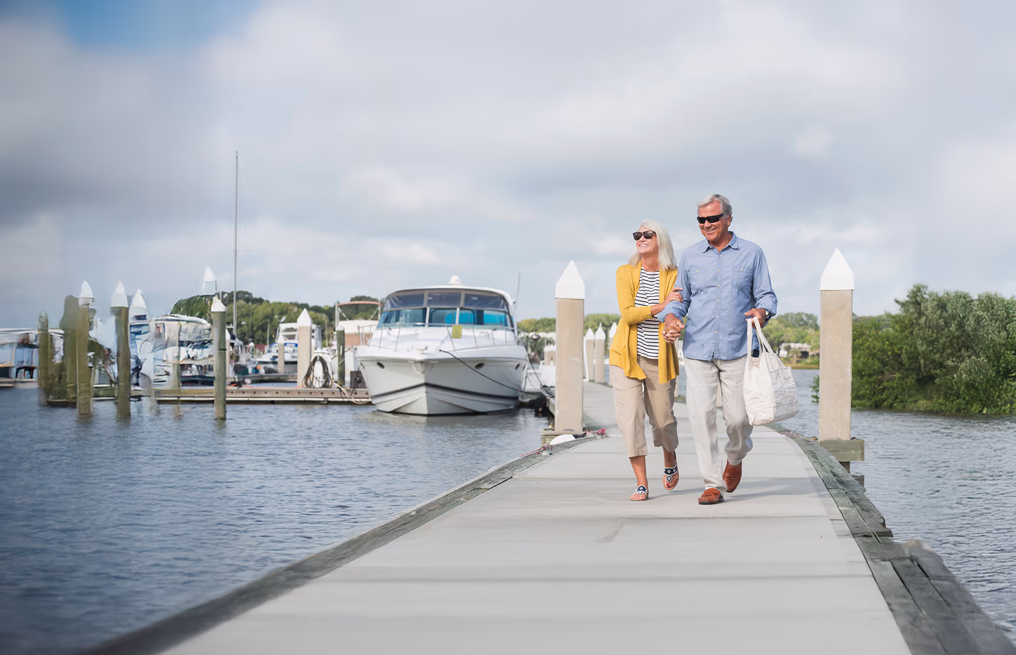 An older couple walks together on a dock near moored boats, smiling and holding a bag.
