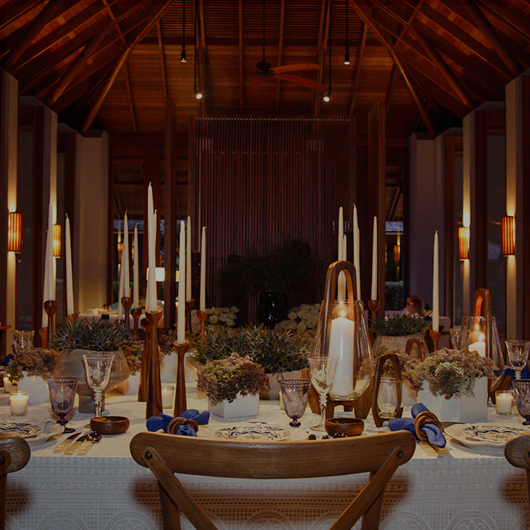 Elegant dining table with white tablecloth, tall white candles in wooden holders, floral centerpieces, and glassware under a wooden vaulted ceiling.