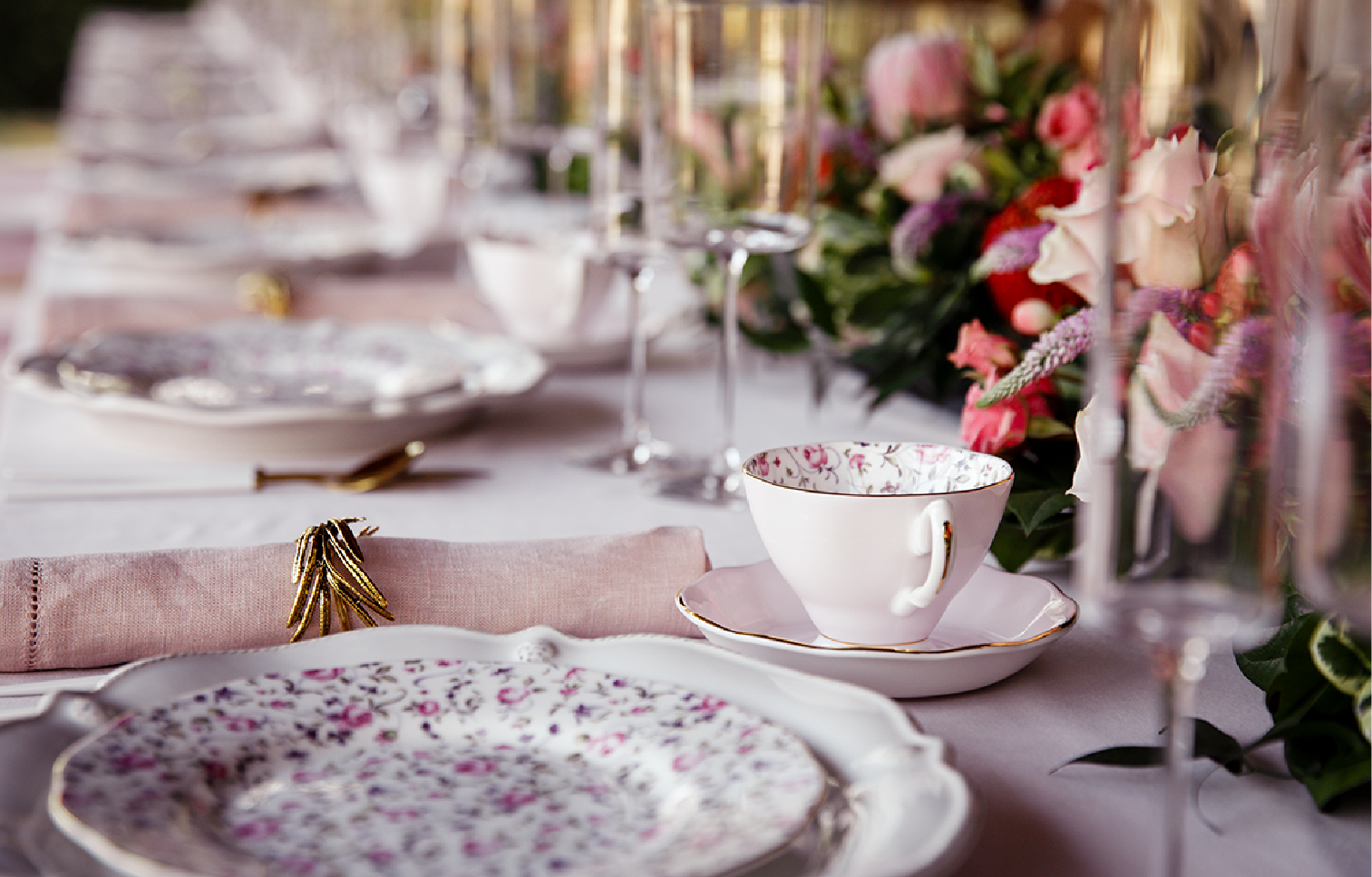 Elegant table setting with floral-patterned china, pink napkin with gold ring, and floral centerpiece.
