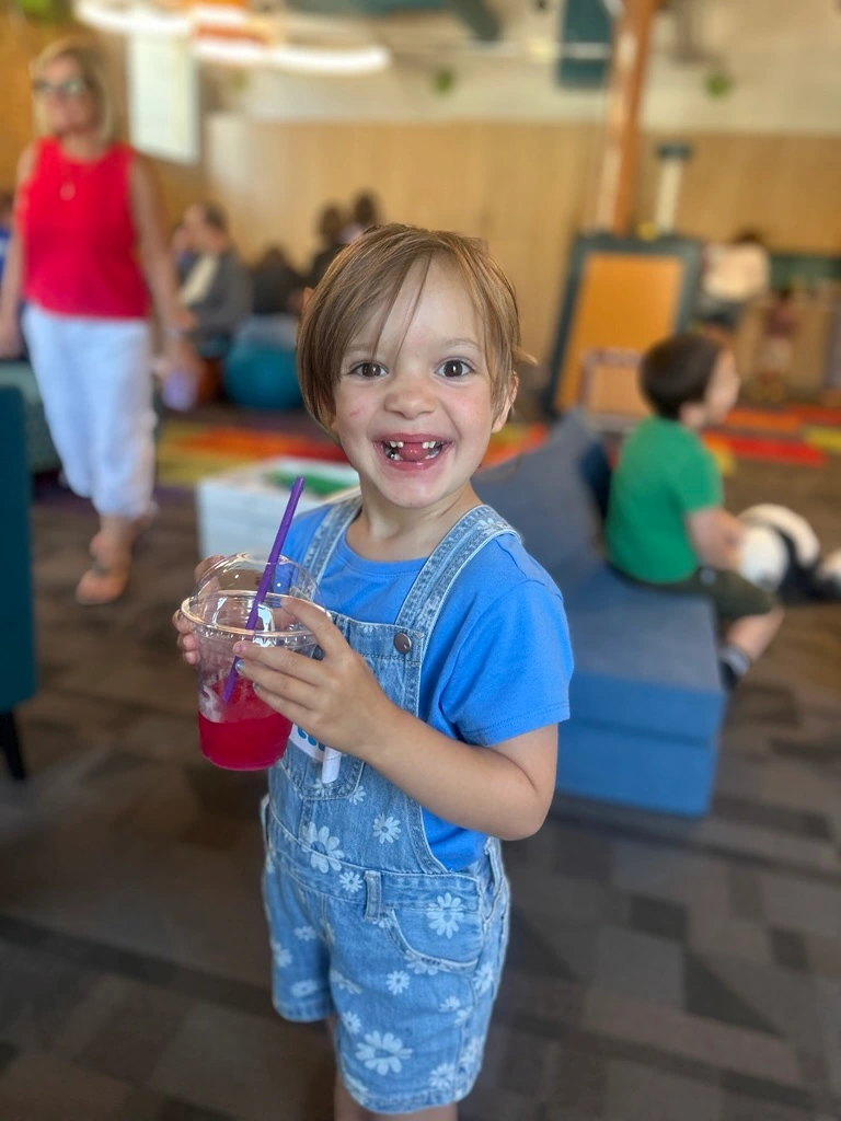 Dream Kid Ella with light brown hair, smiling enthusiastically, holds a pink drink with a straw. Wearing blue floral overalls, in a lively indoor setting.