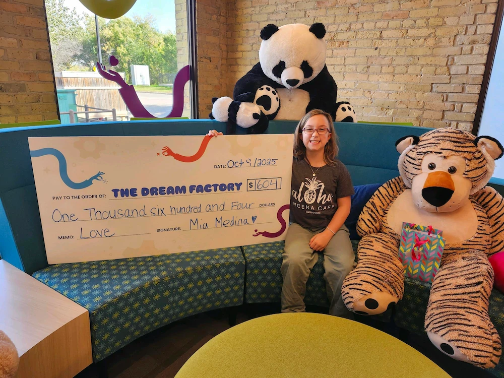 A smiling girl sits between large stuffed panda and tiger toys holding a big check for The Dream Factory worth $1,604.