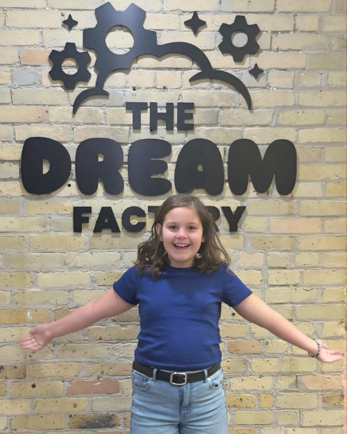 Sophia standing with her arms outstretched in front of The Dream Factory entrance sign.
