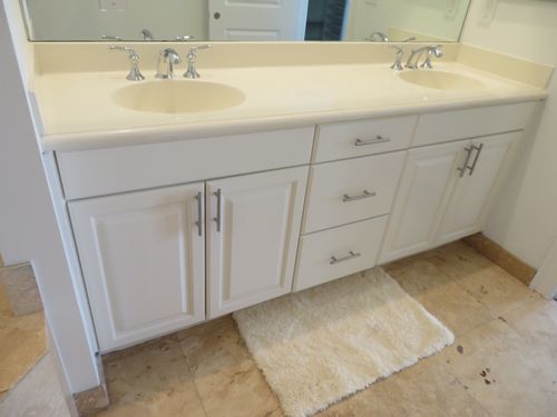 White bathroom vanity with dual sinks, silver faucets, drawers, and a beige rug on tile floor.