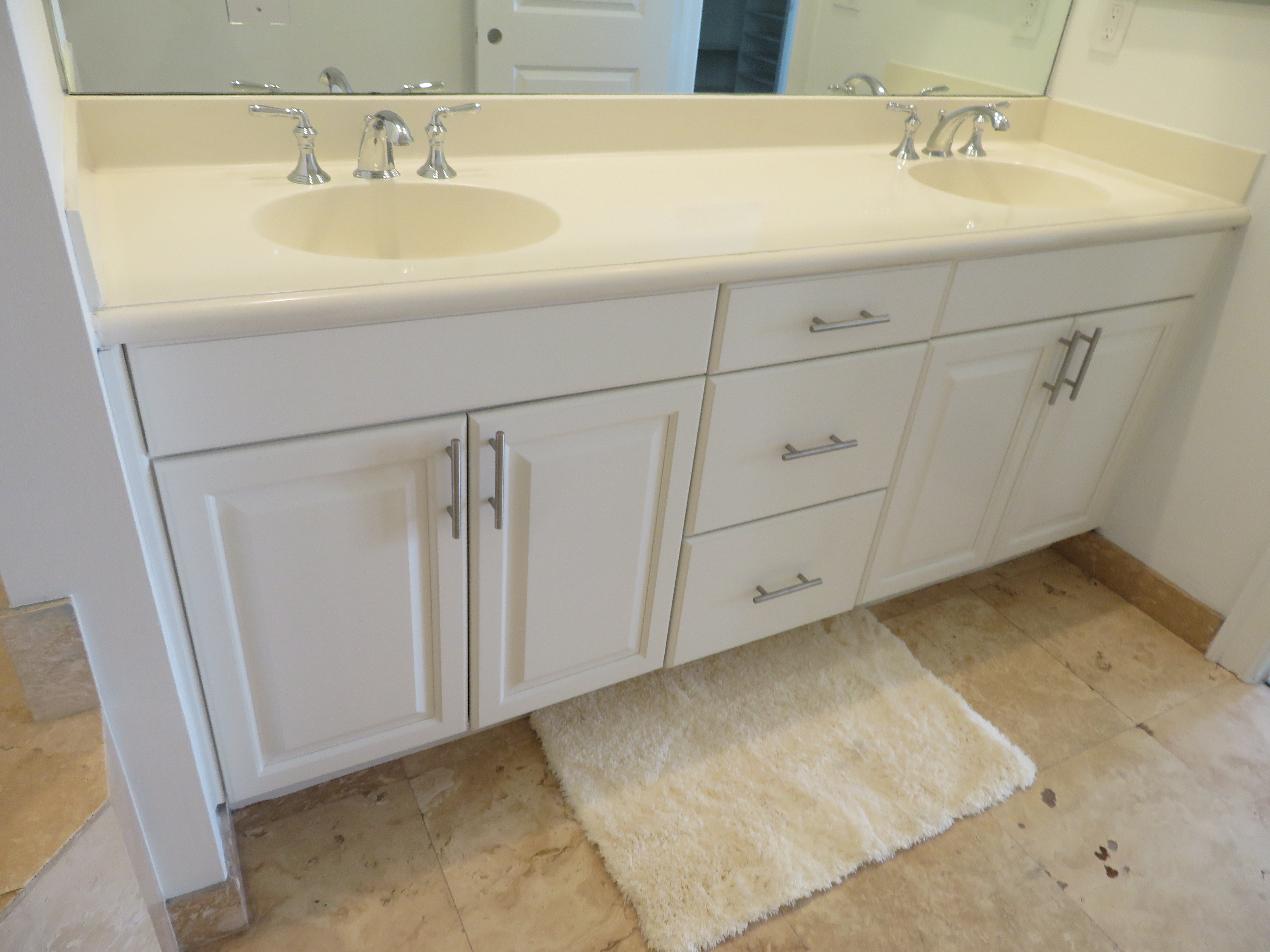 White bathroom vanity with dual sinks, silver faucets, drawers, and a beige rug on tile floor.