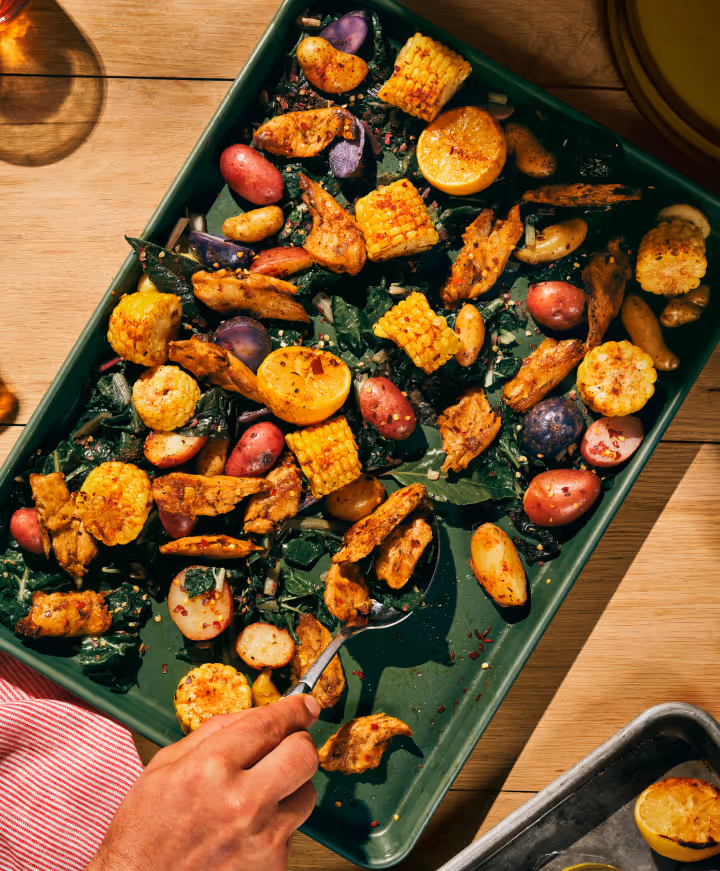 Top-down view of a baking tray with roasted chicken pieces, corn on the cob, purple and red potatoes, kale, and lemon halves, with a hand holding a spoon.