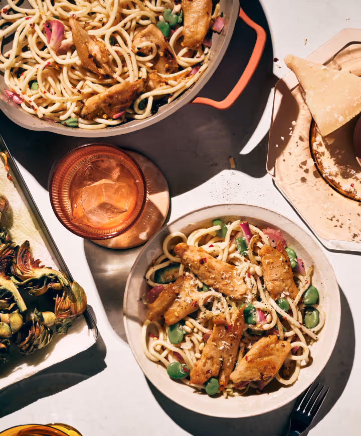 Overhead view of bowls with noodles, grilled chicken pieces, green vegetables, and a glass with an iced drink on a white table.