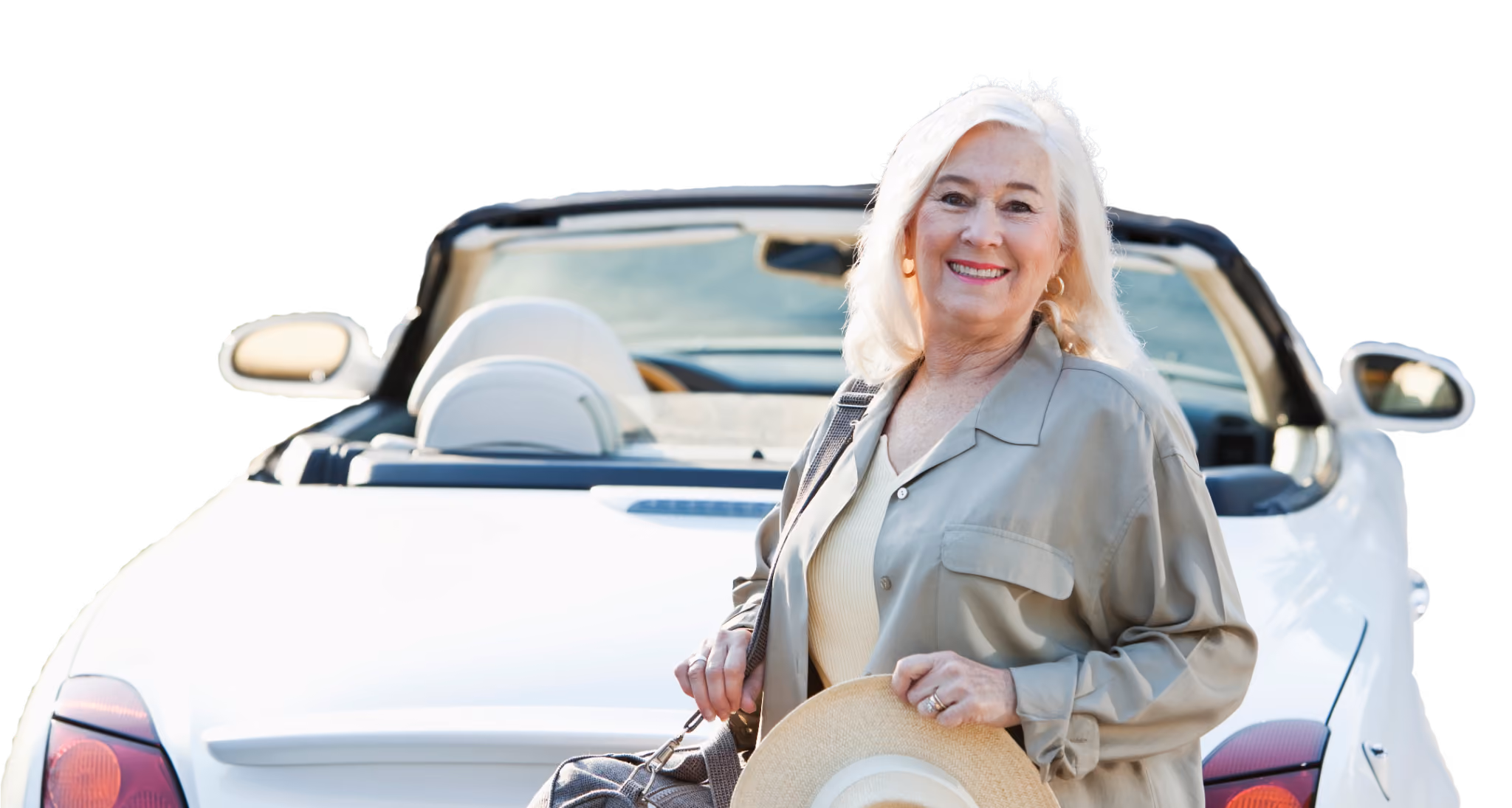 Smiling older woman with white hair standing in front of a white convertible car, holding a straw hat and wearing a light beige outfit.