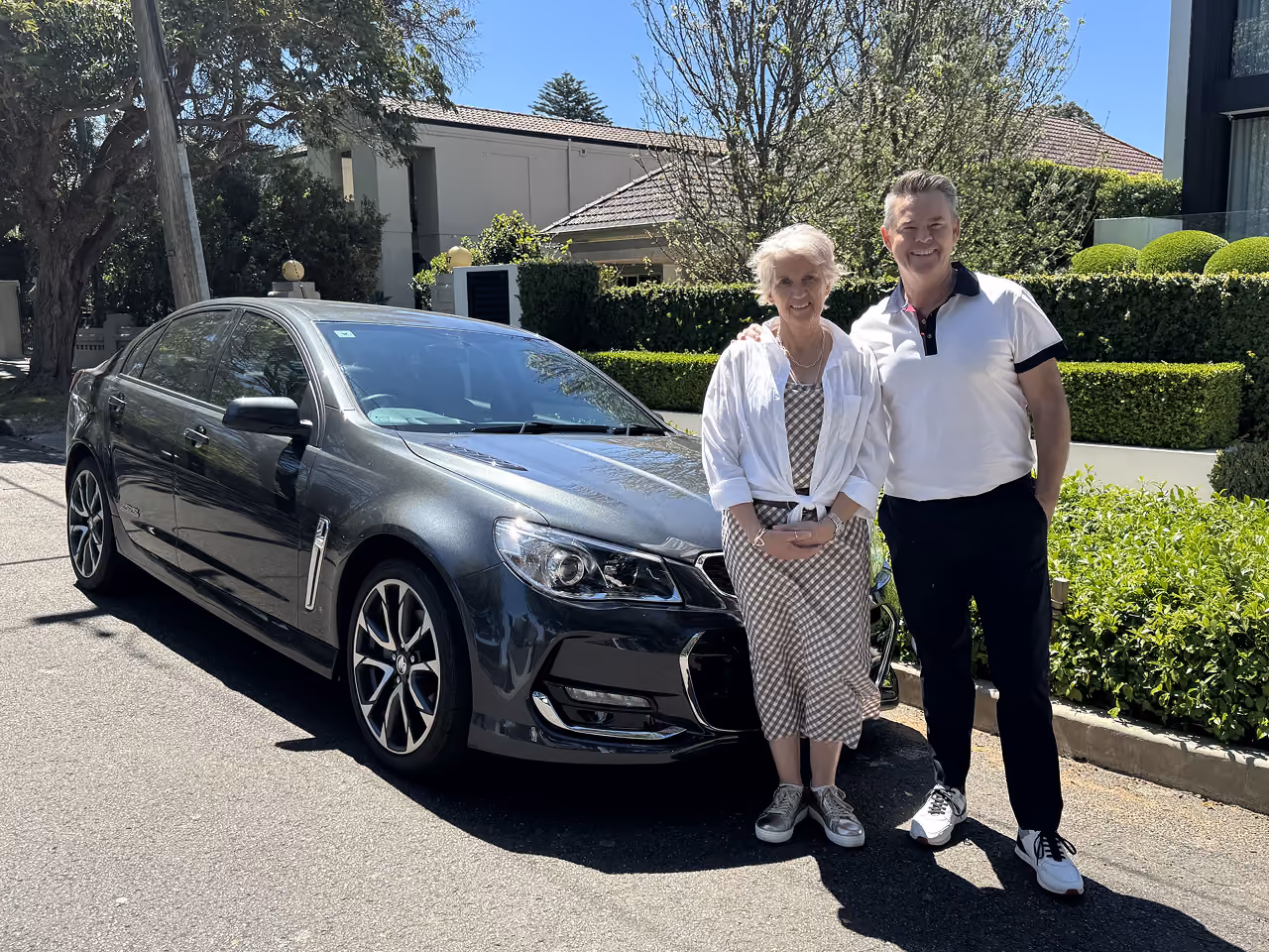 Older man and woman standing together on a sunny residential street beside a dark gray sedan, smiling at the camera, with the woman wearing a patterned dress and light jacket and the man wearing a white polo shirt and dark pants, with well-kept hedges, trees, and houses in the background.