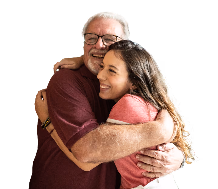 Older man warmly hugging a younger woman likely his daughter inside a home, both smiling, while another woman likely his wife stands nearby smiling in the background.